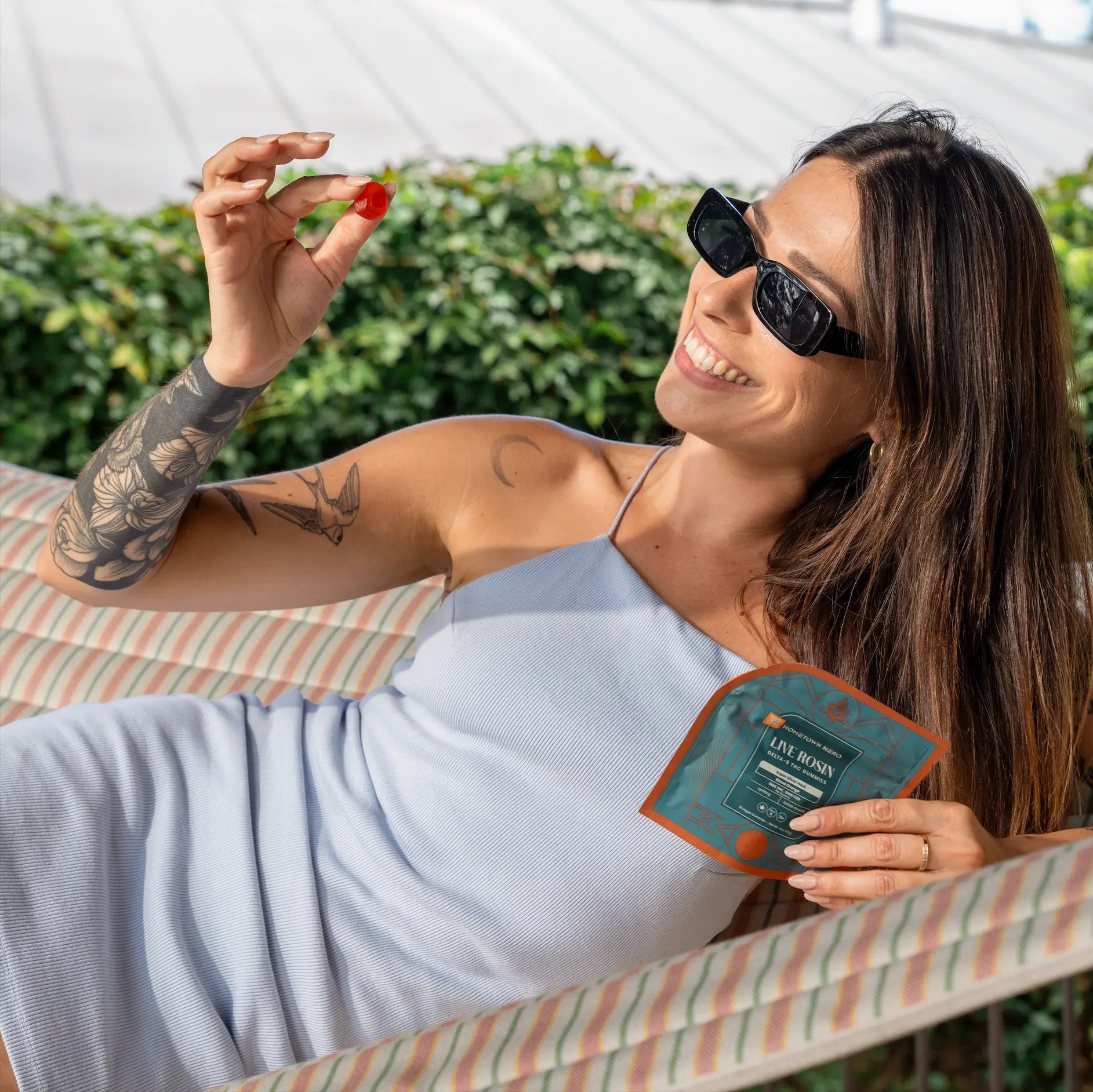 girl wearing sunglasses sitting in a hammock smiling holding a bag of gummies in one hand and a gummy in the other