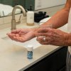 older man holding a pill in one hand and a glass of water in the other by the sink in a bathroom with capsules packaging by the sink